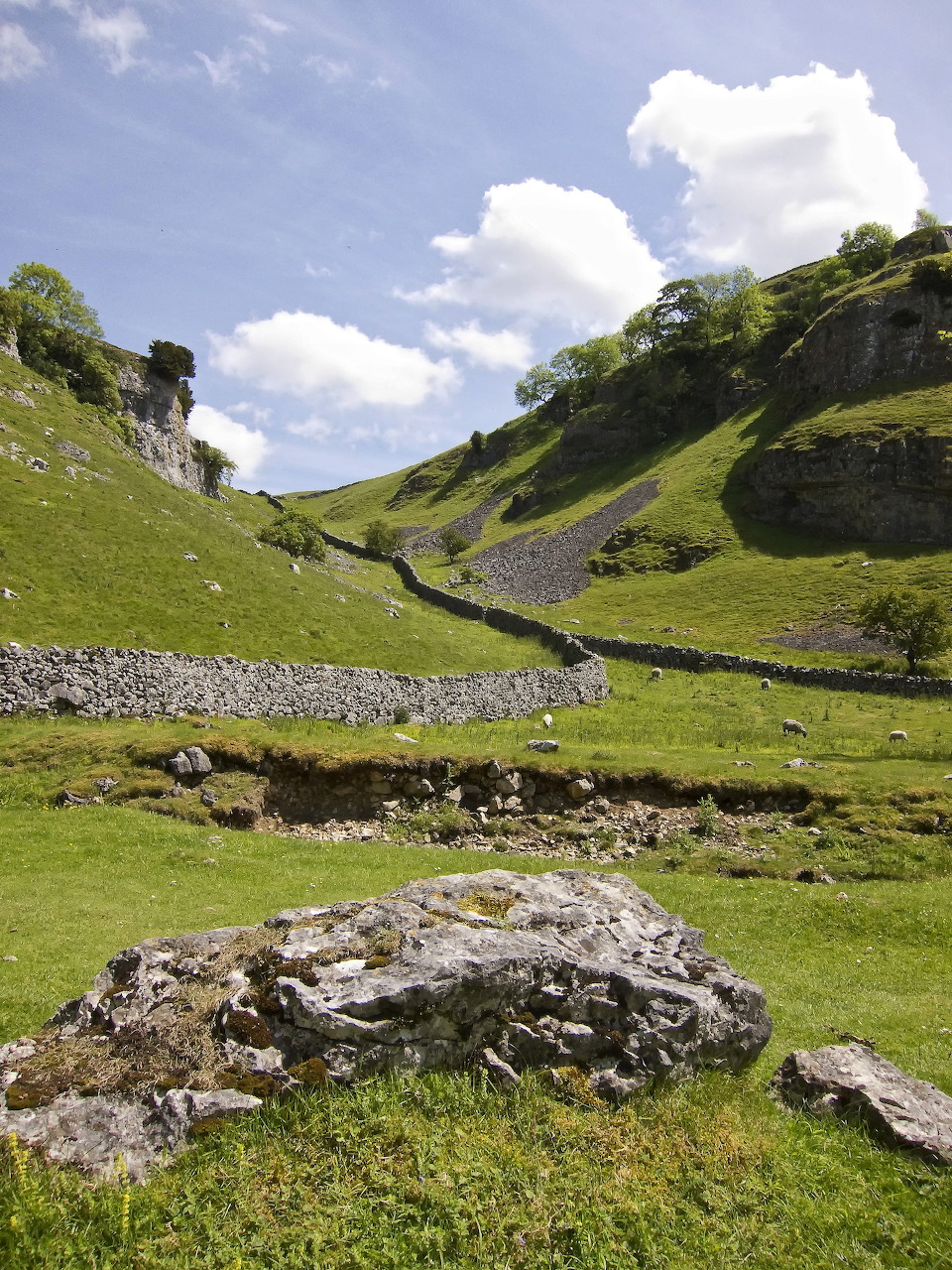 Green upland valley with stone walls, rocky hills and a small stream beneath a blue sky