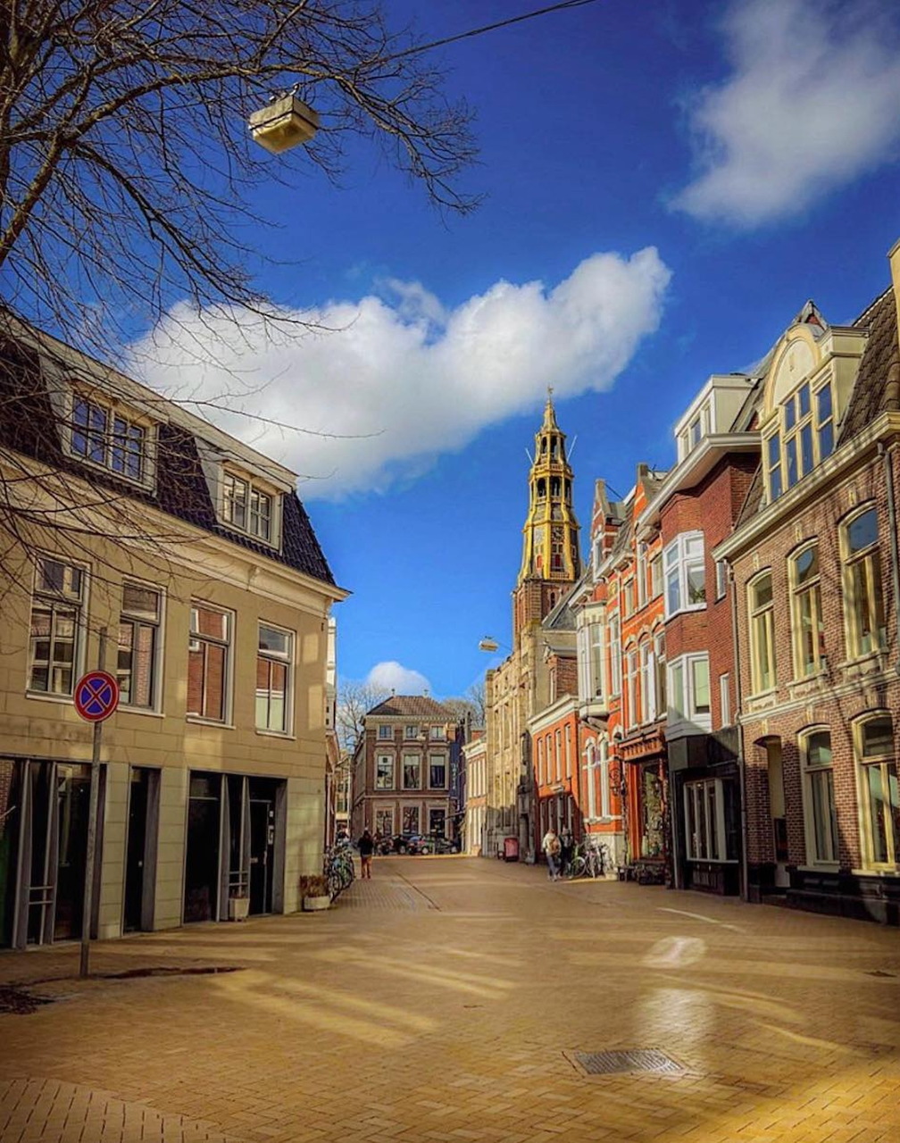 Historic European street with tall old buildings, bare tree branches, and a bright blue sky