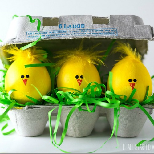 Three yellow Easter chick decorations with orange beaks sitting in egg carton with green shredded paper grass