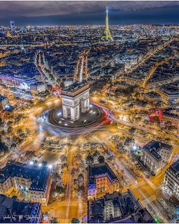 Aerial night view of Paris showing Arc de Triomphe, radiating streets, and illuminated Eiffel Tower in distance