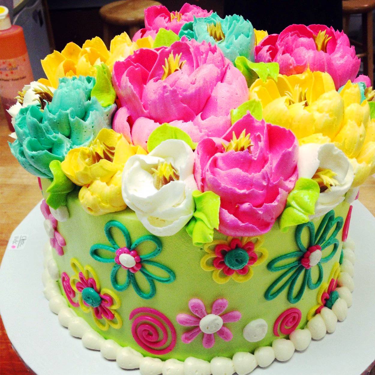 Round cake decorated with bright buttercream flowers in pink, yellow, white, and turquoise on top with colorful flower designs on sides