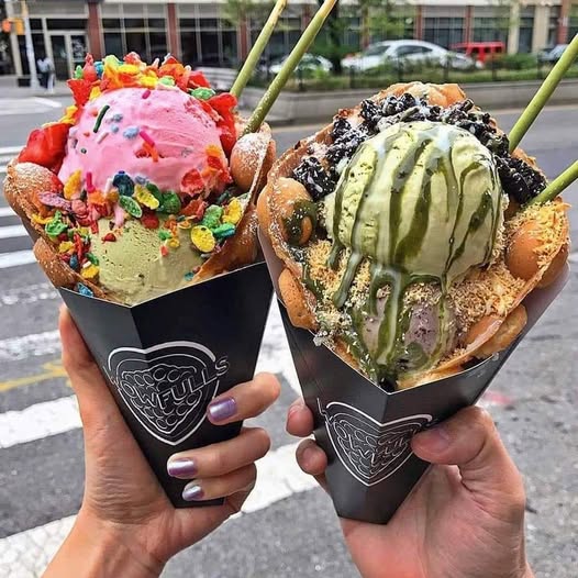Two gelato cones held up, one pink with sprinkles, one green with chocolate drizzle, city street background