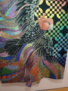 Detailed fabric quilt art showing a cockatoo with colorful stitched feathers and patterned textiles on dark background