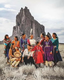 Group of Native American women in traditional dress posing before Shiprock rock formation in desert landscape