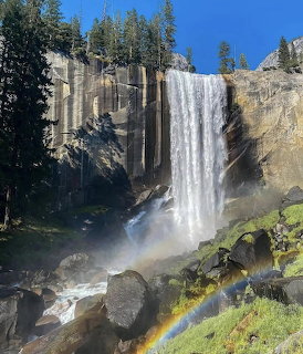 Tall waterfall plunging down rocky cliff with rainbow mist at base, pine trees and blue sky visible