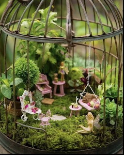 Miniature fairy garden with figurines, swing, and moss inside a metal birdcage surrounded by greenery