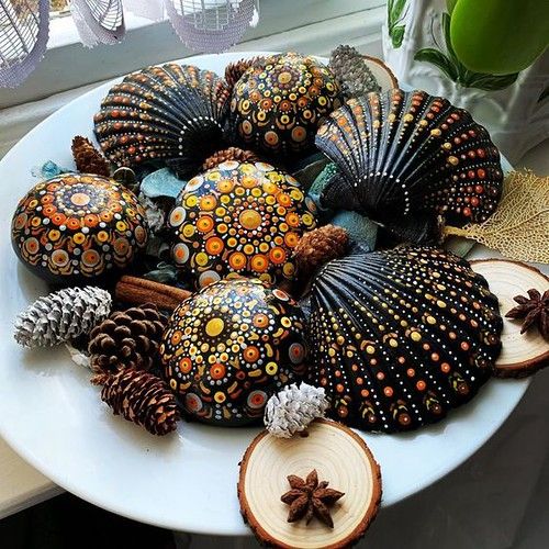 Decorative seashells hand-painted with colorful mandala dot patterns on white plate with pine cones