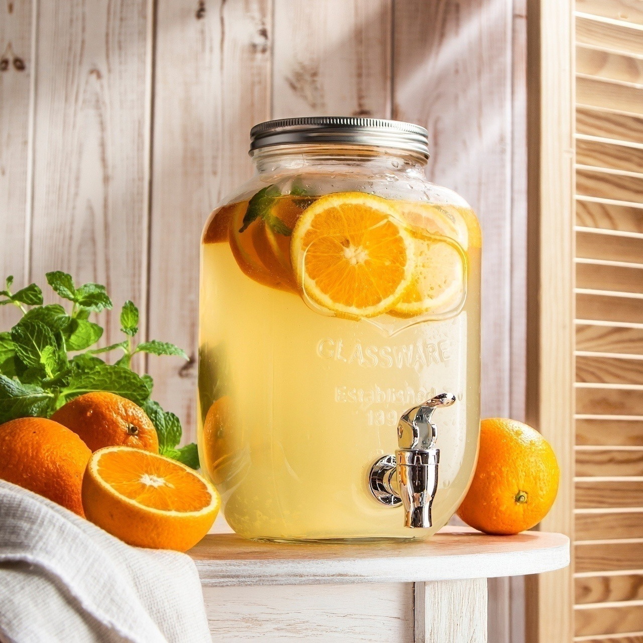 Glass drink dispenser with orange slices in yellow water on white rustic table