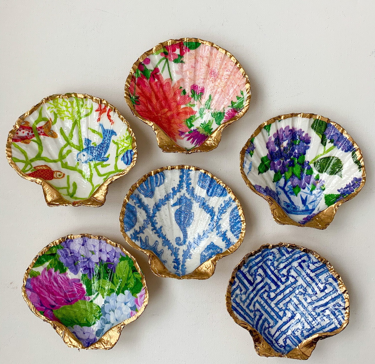 Six hand-painted scallop shells with colorful floral and geometric patterns arranged on beige surface