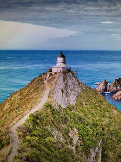 White lighthouse on a green coastal cliff with a winding path, ocean and rocks below, cloudy sky above