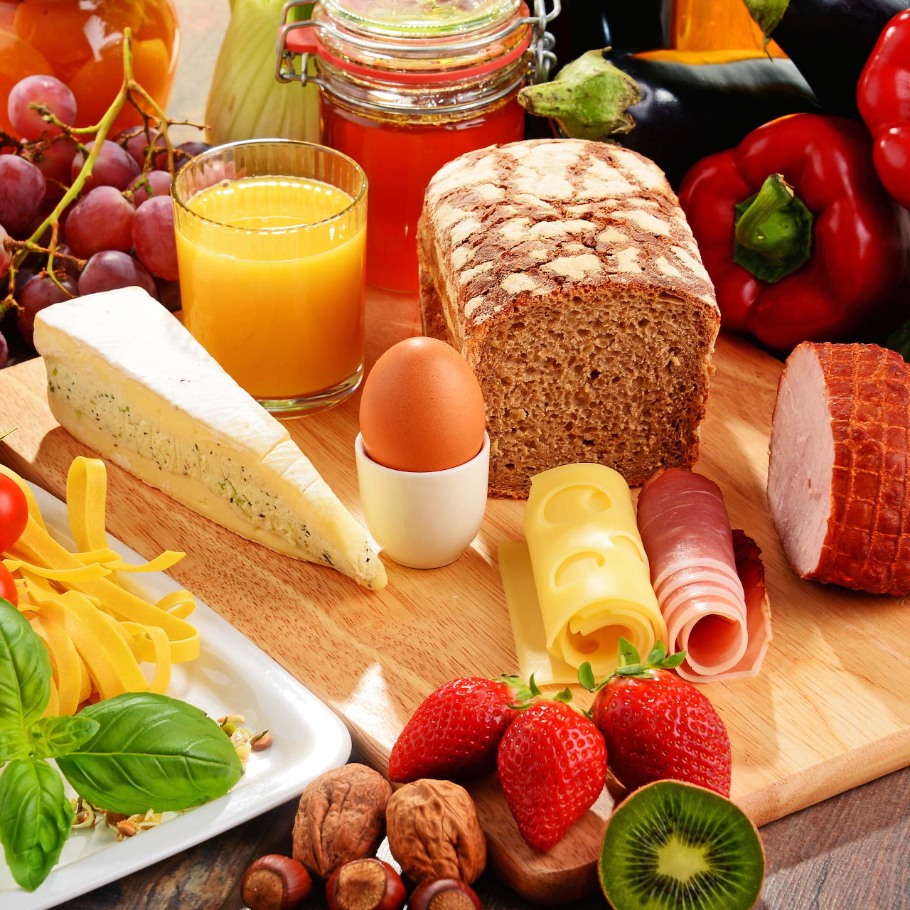 Assorted breakfast foods including bread, cheese, meats, fruits, vegetables, and beverages on wooden cutting board