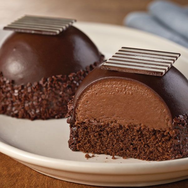 Two chocolate dome desserts with ganache coating and decorative chocolate squares on white plate
