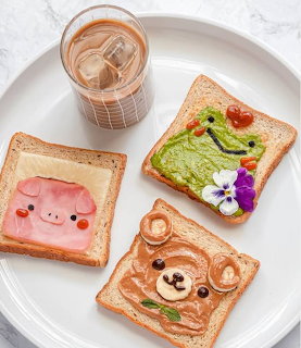 Four slices of toast decorated with cute animal faces using spreads, fruits, and edible flowers on a white plate