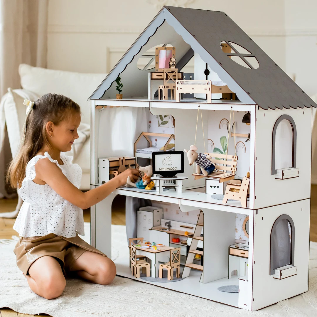 Young girl kneeling beside a large white dollhouse with miniature furniture indoors