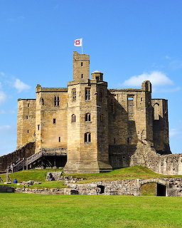 Warkworth Castle, a medieval stone fortress with central tower flying a flag, set against blue sky and green grass