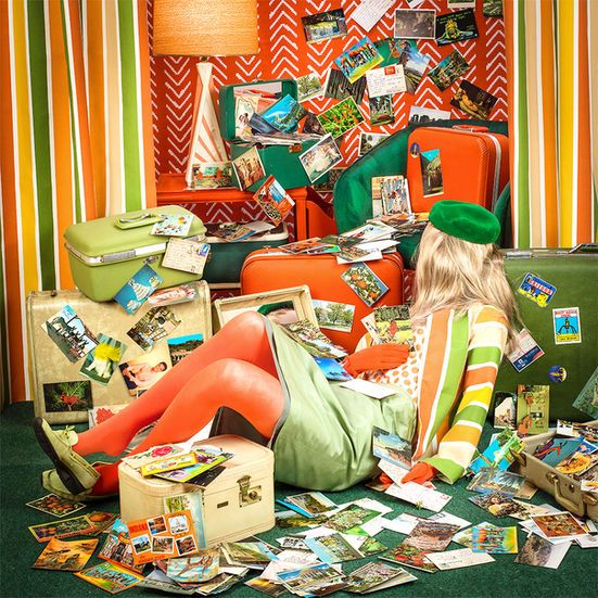Blonde woman in retro clothes sitting among colorful suitcases and postcards in a striped room