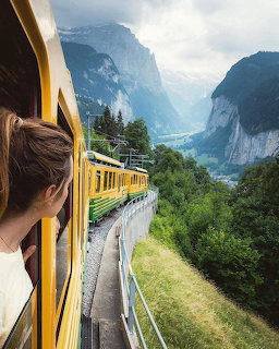 Woman leaning out of a yellow train window overlooking a green Swiss valley with mountains and cliffs