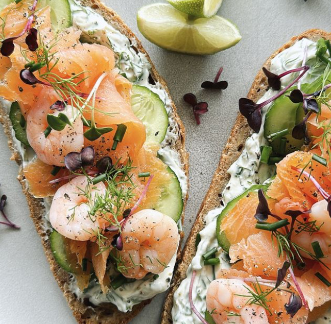 Two pieces of toast topped with shrimp, avocado, salmon, cream cheese and herbs