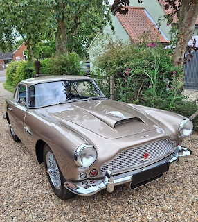 Champagne gold Aston Martin DB4 classic car parked on gravel in front of house with trees