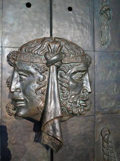 Bronze relief sculpture of a two-faced classical head on a stone wall