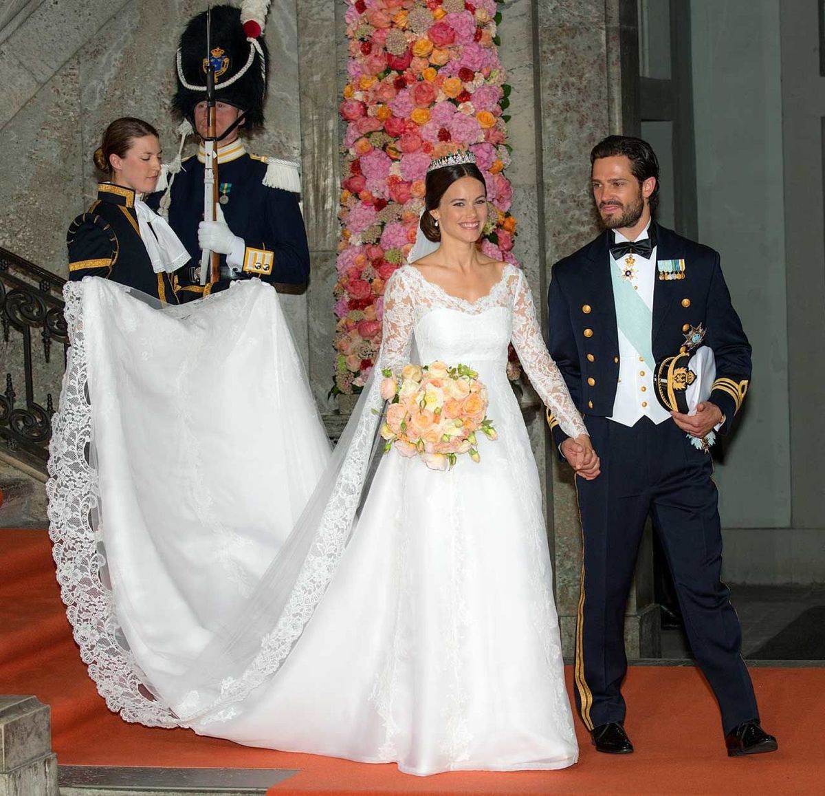 Bride and groom in formal wedding attire walking on red carpet with attendants holding dress train