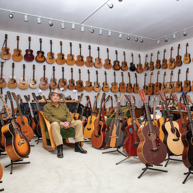 Man sitting in room filled with numerous acoustic guitars on walls and floor