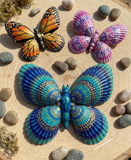 Three butterflies made from painted seashells in orange, purple, and turquoise blue on wooden surface with smooth stones