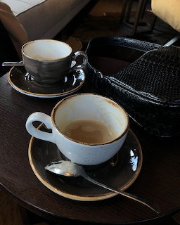 Two white coffee cups on saucers on a dark wooden table with a spoon and a black woven bag