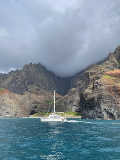 White catamaran sailing near towering Na Pali Coast cliffs in Kauai with clouds covering mountain peaks