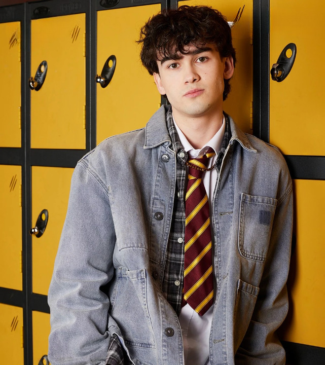 Young man in school uniform with striped tie and grey denim jacket standing in front of yellow lockers