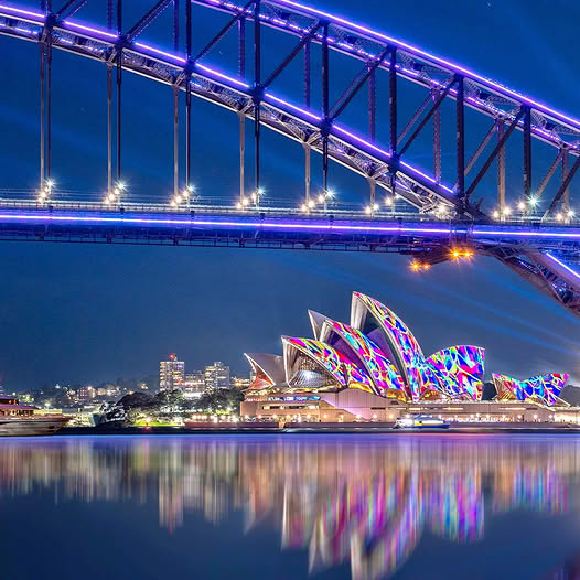 Colorful Sydney Opera House at night beneath a bridge, reflected in the harbor