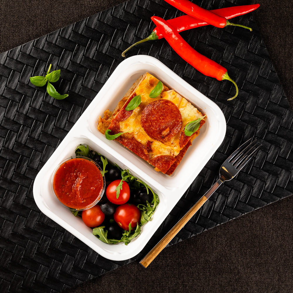 Lasagna in white takeout box with tomatoes, greens, sauce, chopsticks and red chilies on dark woven mat