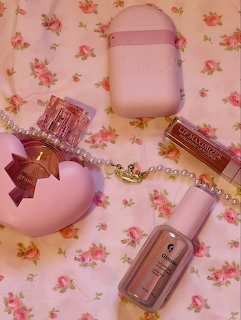 Pink beauty products arranged on a floral fabric including perfume bottle, lip product, and skincare