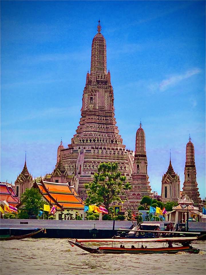 Tall ornate temple tower with intricate carvings and smaller spires beside a river with traditional boats