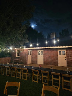 Outdoor banquet setup at night with wooden chairs, brick building, and string lights under moon