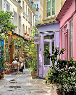 Narrow colorful Paris alley with pink and purple buildings, green plants, and a café terrace