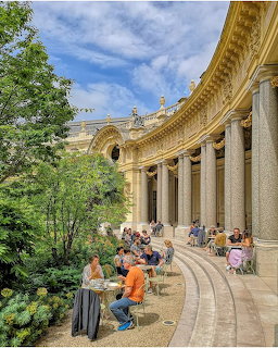 People dining at an outdoor cafe beneath a grand colonnade with classical columns and lush greenery