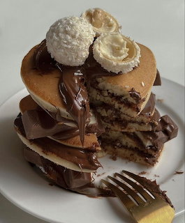Stack of pancakes covered in chocolate sauce topped with cream and coconut balls on a white plate