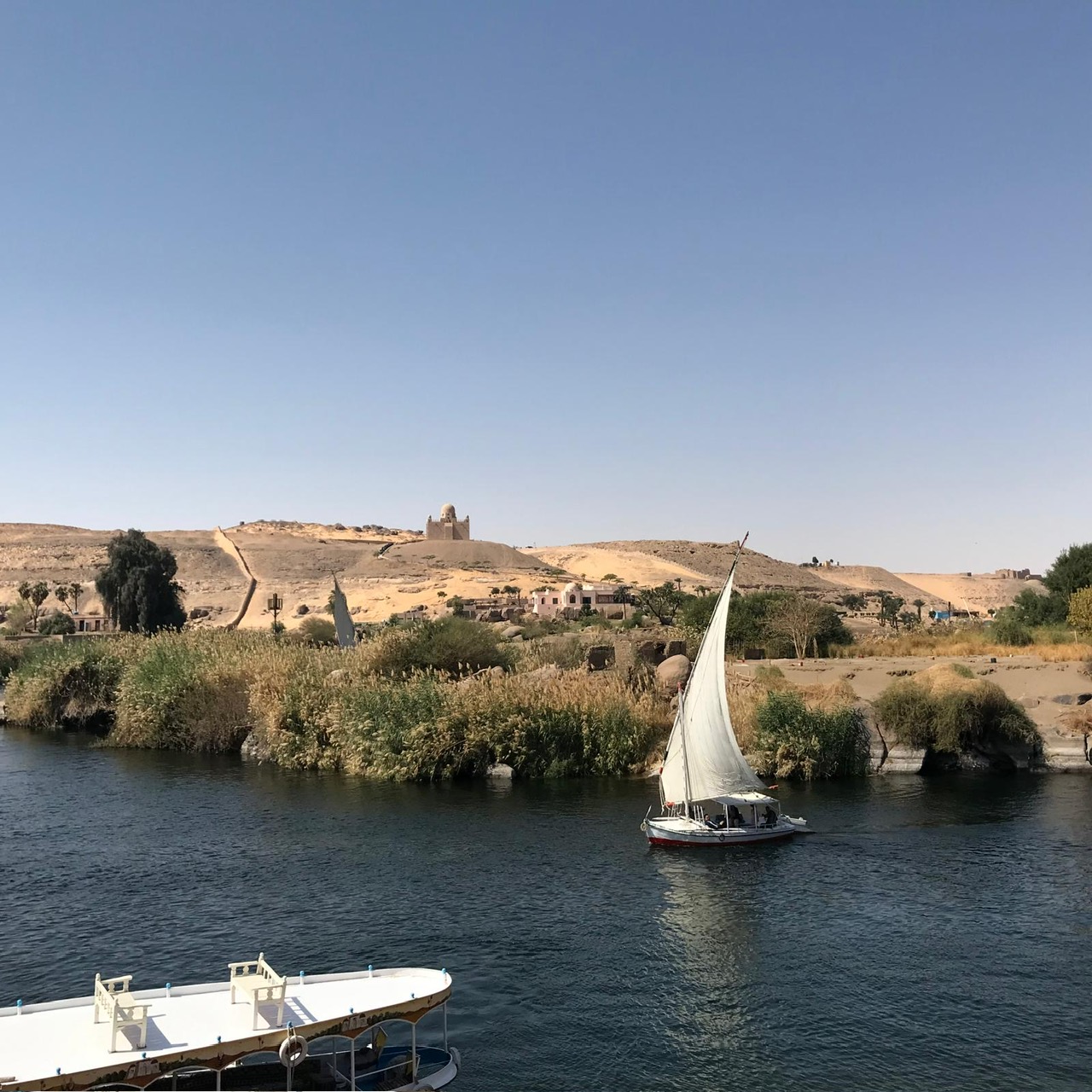 Felucca sailboat on the Nile River with green riverbank vegetation and desert hills with ruins behind