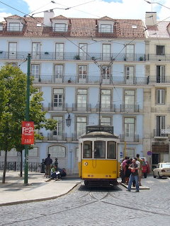 Yellow historic tram on a cobblestone street in Lisbon, Portugal, with a white multi-story building behind