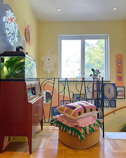 Colorful eclectic living room with yellow walls, wicker chair, fish tank on red cabinet, and neon wall art