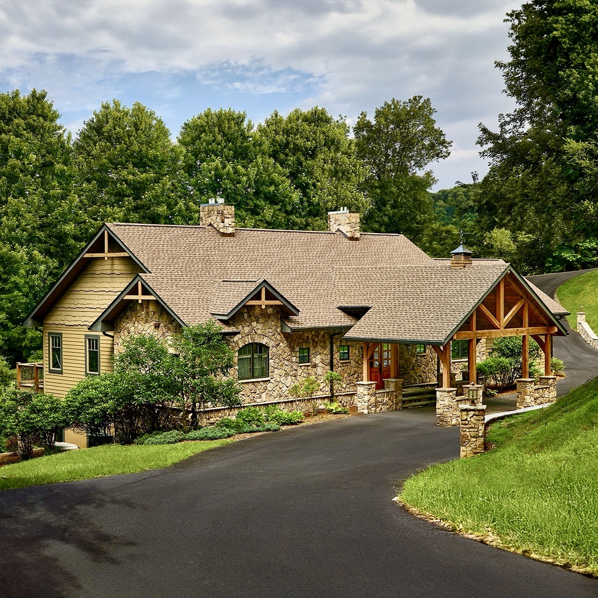 Craftsman home with stone pillars and peaked rooflines on wooded hillside with paved driveway
