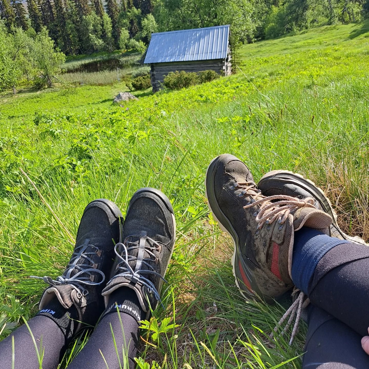 Two pairs of hiking boots on green grass with mountain cabin behind