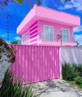 Bright pink two-story house with white railings, blue sky, and plants in front