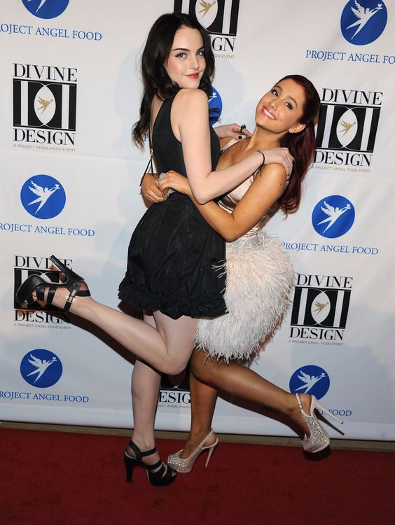 Two young women posing on a red carpet at a charity event, one in a black dress and one in a white feathered dress.