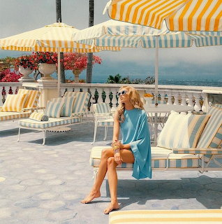 Blonde woman sitting on a seaside terrace with striped umbrellas, lounge chairs, and ocean view