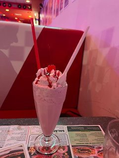 Pink milkshake with whipped cream and cherry in a tall glass at a retro diner with red booths