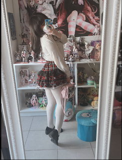 Young woman in plaid skirt and white tights taking a mirror selfie with anime figurines on shelves behind her