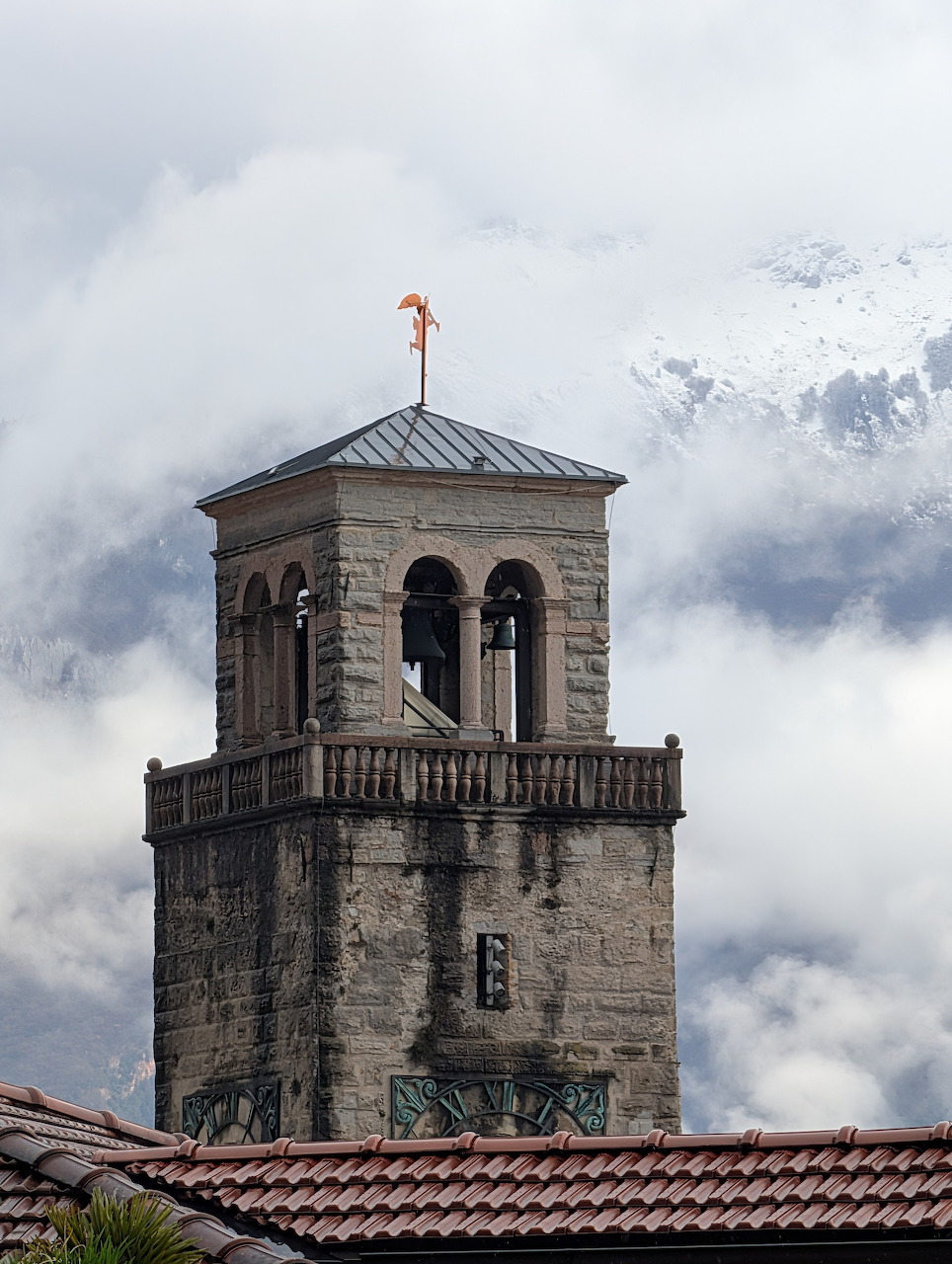 Stone bell tower above a red tiled roof with clouds and mountains behind