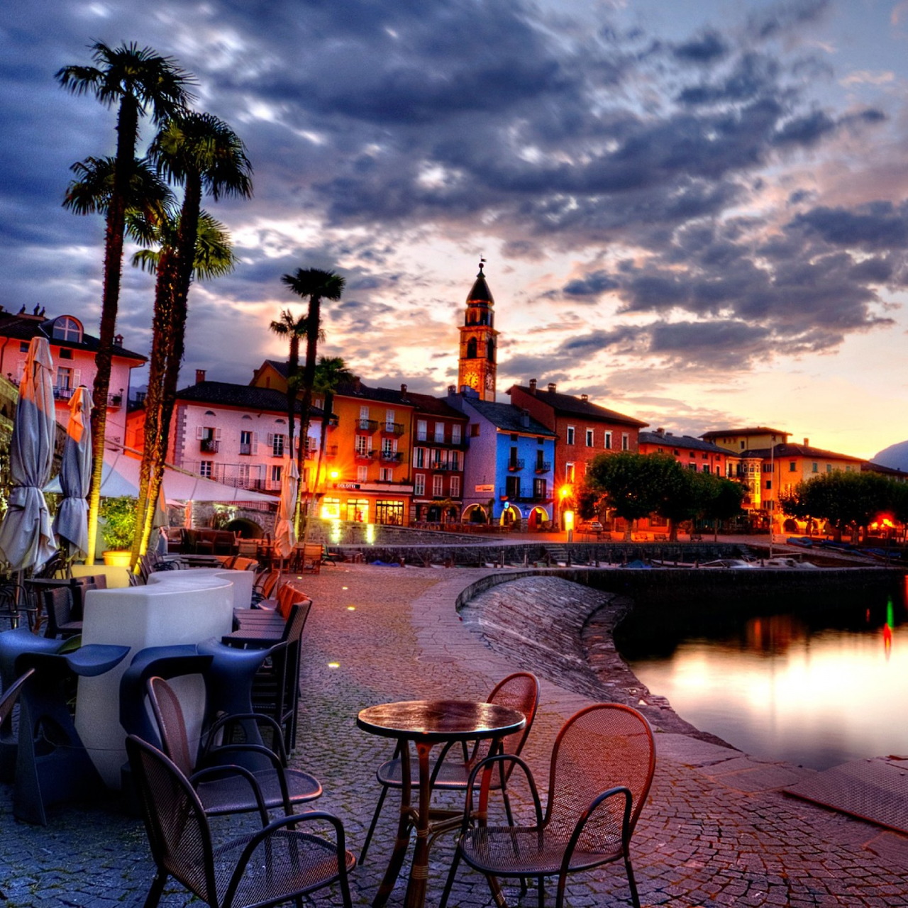 Sunset view of a European waterfront town with colorful buildings, a church tower, palm trees, and café tables by the water